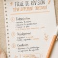 Une femme souriante crée une fiche de révision sur sa table de travail, entourée de fournitures d'étude.